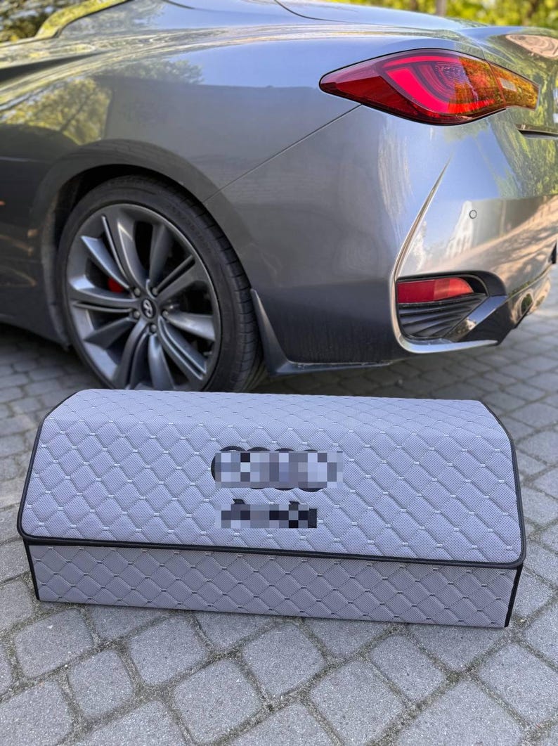 May include: A gray car trunk organizer with a quilted pattern. The organizer is rectangular with a black trim and is positioned in front of a gray car. The car has a red taillight.