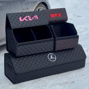 May include: Black quilted car trunk organizers with pink and red text accents. The organizers are stacked, featuring multiple compartments and a logo. The design includes a diamond pattern.
