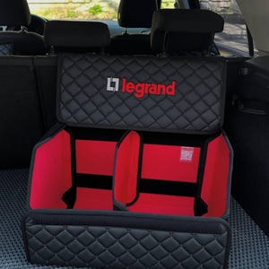 以下が含まれることがあります： 「Legrand」のロゴが入った黒と赤の車のトランクオーガナイザー。オーガナイザーの外側はキルティング模様で、赤い内装の2つのコンパートメントがあります。車の後部に収まるように設計されています。