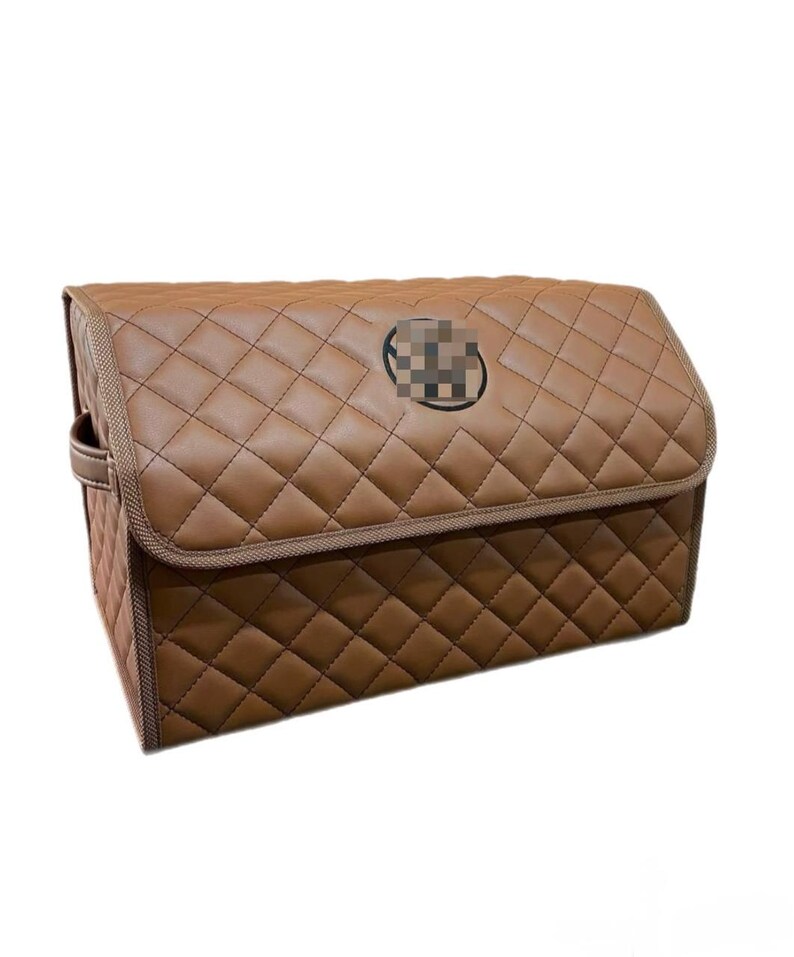 May include: Brown quilted car organizer with a diamond pattern. The organizer has a rectangular shape with a lid and a handle on the side. A circular emblem is on the front.
