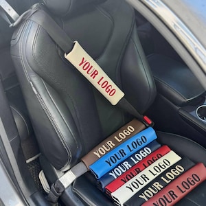 以下が含まれることがあります： 赤い「YOUR LOGO」の文字が入った車のシートベルトカバー。茶色、青、ネイビー、赤、白、黒、バーガンディなど、さまざまな色のカバーが黒い革張りの車のシートに並んでいます。車の内装も写っています。