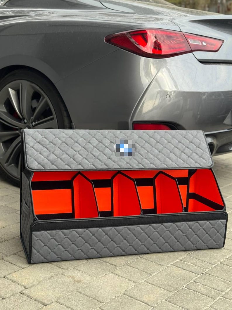 Może przedstawiać: Szary pikowany organizer do bagażnika samochodowego z czarnym wykończeniem i pomarańczowymi przegrodami wewnętrznymi. Posiada logo BMW na pokrywie. Zaprojektowany, aby utrzymać bagażnik samochodu w porządku.