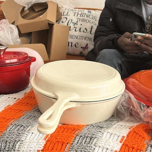Könnte beinhalten: Eine cremefarbene gusseiserne Bratpfanne mit Griff liegt auf einer bunten orangefarbenen, grauen und weißen Strickdecke. Ein roter Dutch Oven und ein orangefarbener Deckel sind ebenfalls sichtbar. Der Hintergrund umfasst Pappkartons und ein Kissen mit Text.