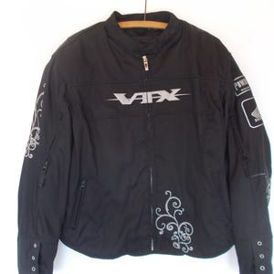 May include: Black motorcycle jacket with the word "VAFX" in silver across the chest. The jacket has silver decorative swirls on the sleeves and front. A "HONDA" patch is on the right sleeve. The jacket has a zipper closure and multiple pockets.