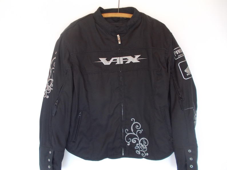 May include: Black motorcycle jacket with the word "VAFX" in silver across the chest. The jacket has silver decorative swirls on the sleeves and front. A "HONDA" patch is on the right sleeve. The jacket has a zipper closure and multiple pockets.