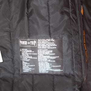 May include: Black quilted nylon jacket interior with a Power-Trip label. The label includes care instructions and fabric composition details. The jacket has a zipper and the text "LESS BRAKE MORE THROTTLE" is visible.