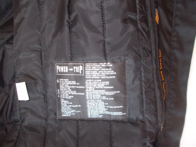 May include: Black quilted nylon jacket interior with a Power-Trip label. The label includes care instructions and fabric composition details. The jacket has a zipper and the text "LESS BRAKE MORE THROTTLE" is visible.