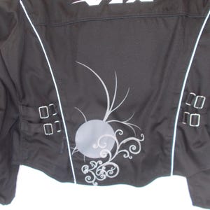 May include: Black motorcycle jacket with white accents and decorative silver embellishments. Features a gray circular design with swirling patterns on the back. Includes silver buckles and a white logo at the top.