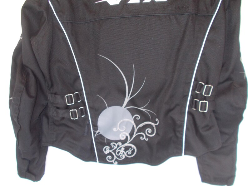 May include: Black motorcycle jacket with white accents and decorative silver embellishments. Features a gray circular design with swirling patterns on the back. Includes silver buckles and a white logo at the top.