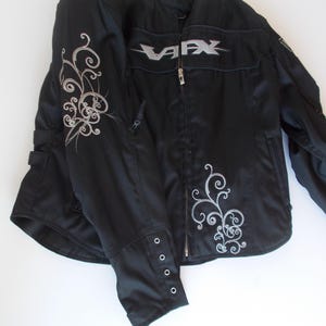 May include: Black motorcycle jacket with silver accents. The jacket features a silver "VFX" logo on the chest and decorative silver swirls on the sleeves and front. It has a zipper closure and buttoned cuffs. A practical and stylish outerwear option.