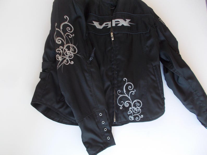 May include: Black motorcycle jacket with silver accents. The jacket features a silver "VFX" logo on the chest and decorative silver swirls on the sleeves and front. It has a zipper closure and buttoned cuffs. A practical and stylish outerwear option.