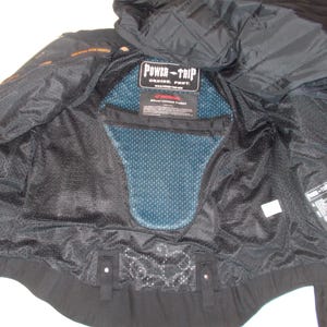 May include: Black motorcycle jacket interior with a blue mesh back protector. The jacket has a quilted hood and the "POWER-TRIP" logo is visible. The interior is made of black mesh and fabric.