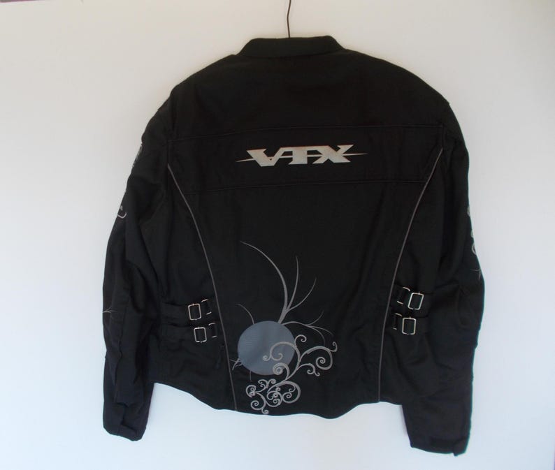 May include: Black motorcycle jacket with the text "VFX" in silver across the back. The jacket has silver accents, a decorative design, and two sets of buckles on the lower back.