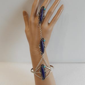 May include: A silver hand chain with a ring, a bracelet, and two connecting chains. The ring, bracelet, and chain links feature oval, dark blue stones with turquoise accents. The jewelry is displayed on a mannequin hand.
