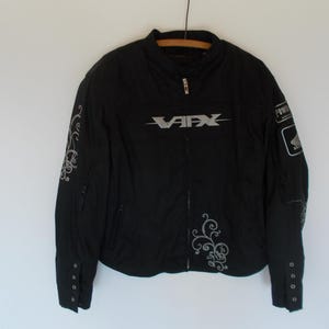 May include: Black motorcycle jacket with silver accents. Features the text "VAFX" across the chest and decorative swirls on the sleeves. Includes a "POWER" patch and a Honda logo on the right sleeve. The jacket has a zipper closure and buttoned cuffs.