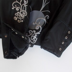 May include: Black jacket with silver embroidery and decorative accents. The jacket features a buttoned cuff and a detailed floral design on the front. The sleeves have buttoned cuffs and the jacket is made of a textured material.