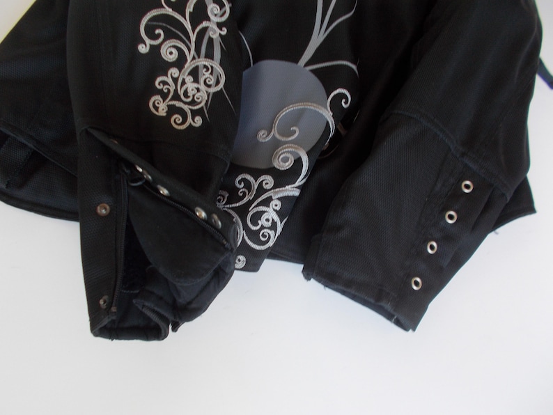 May include: Black jacket with silver embroidery and decorative accents. The jacket features a buttoned cuff and a detailed floral design on the front. The sleeves have buttoned cuffs and the jacket is made of a textured material.