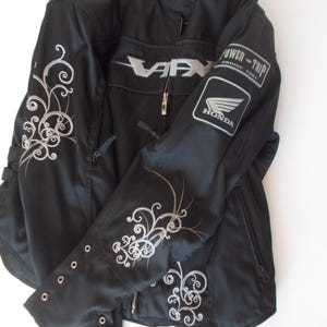 May include: Black motorcycle jacket with silver floral embroidery on the sleeves and shoulders. Features a zippered front, a Honda logo patch, and the word "VARX" in silver. The jacket has a stand-up collar and multiple pockets.