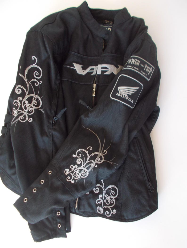 May include: Black motorcycle jacket with silver floral embroidery on the sleeves and shoulders. Features a zippered front, a Honda logo patch, and the word "VARX" in silver. The jacket has a stand-up collar and multiple pockets.