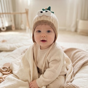 May include: A baby wearing a beige knit hat with large cartoon eyes and a green fin, paired with a matching beige sweater and pants. The baby is wrapped in a cream-colored blanket, sitting on a cream-colored mat.