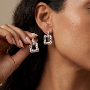 May include: Silver-toned square earrings with a textured, hammered metal finish. The earrings have a rectangular shape with a smaller rectangular top piece. The earrings are shown on a person's ear.