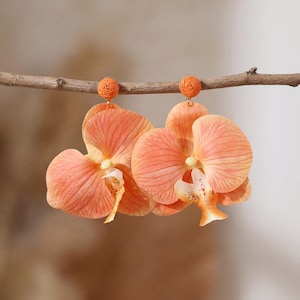 May include: Peach-colored orchid earrings with detailed petals and a textured, circular top. The earrings are suspended from a thin, brown branch against a soft, blurred background. The earrings' design mimics the natural beauty of orchids.