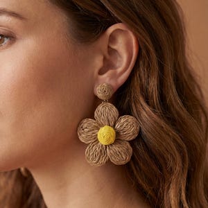 May include: Close-up of a pair of large flower-shaped earrings. The earrings are made of woven, light brown material with a yellow center. The earrings are attached to a small, round stud at the top.