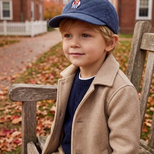 May include: A young child wearing a navy blue quilted baseball cap with a bear emblem, a tan coat, a navy blue sweater, and khaki pants. The child is sitting on a wooden bench outdoors.