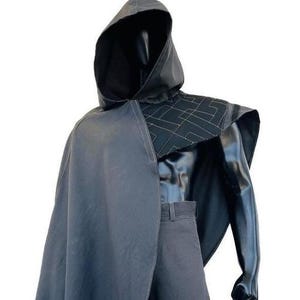 May include: A dark gray hooded cloak and matching pants on a mannequin. The cloak has a cape-like design with a geometric pattern on the shoulders. The outfit is a solid color, with a sleek, modern aesthetic.