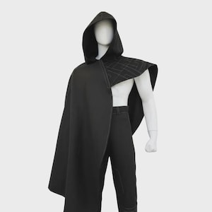 Puede incluir: Una capa negra con capucha y diseño geométrico en los hombros. La capa se combina con pantalones negros. El conjunto se exhibe en un maniquí sobre un fondo blanco. La capa tiene un diseño largo y fluido.