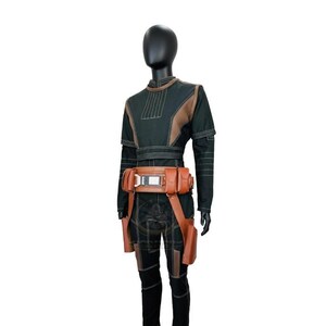 May include: A dark green long-sleeved jumpsuit featuring textured brown panels on the chest and legs, accented with white stitching. A tan utility belt with multiple pouches and a central metallic buckle is worn around the waist of the garment.