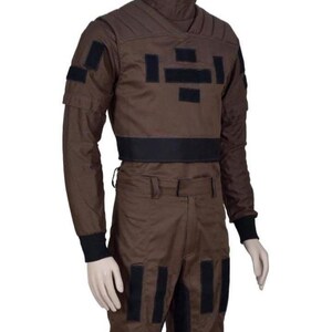 May include: A brown jumpsuit with black accents, displayed on a mannequin. The outfit features a high collar, long sleeves, and multiple black rectangular patches. The pants have pockets and black patches. The cuffs are black.