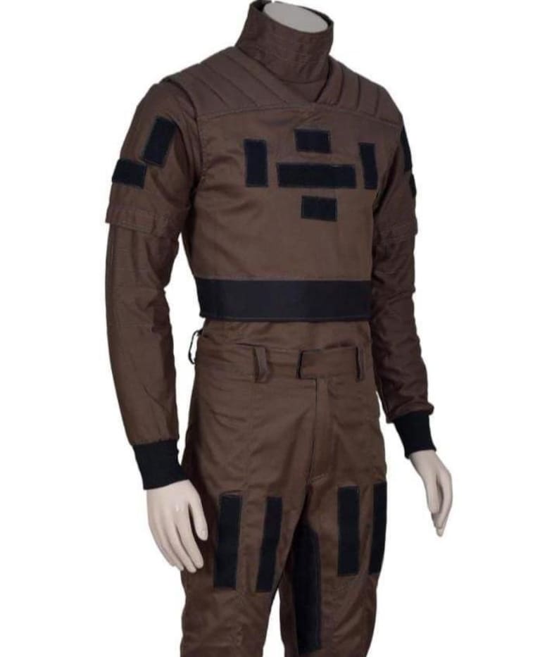 May include: A brown jumpsuit with black accents, displayed on a mannequin. The outfit features a high collar, long sleeves, and multiple black rectangular patches. The pants have pockets and black patches. The cuffs are black.