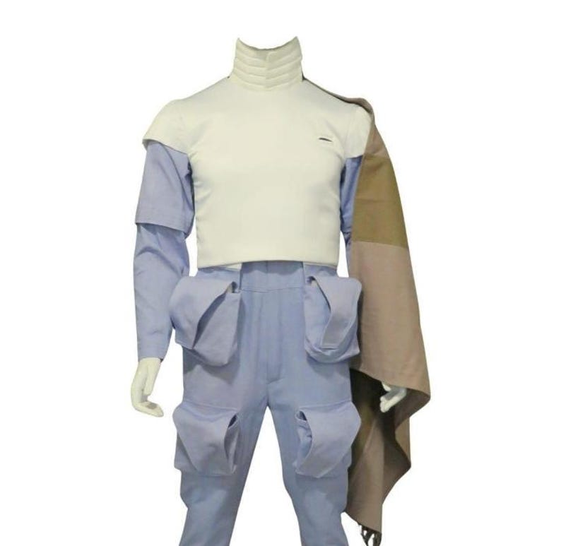 May include: A light blue jumpsuit featuring a white high-neck bodice, long sleeves, and four large cargo-style pockets on the legs. A tan and brown color-blocked fabric panel drapes from the right shoulder, extending down the side of the garment.