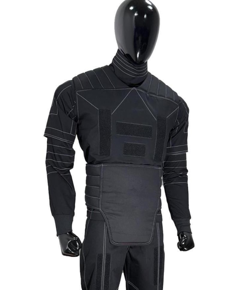 May include: Black tactical suit displayed on a mannequin. The suit features a high collar, long sleeves, and reinforced panels with white stitching. The design includes multiple hook-and-loop fastener patches and a padded lower torso section.