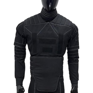 May include: Black tactical vest with white stitching details, displayed on a mannequin. The vest features multiple panels, hook-and-loop closures, and long sleeves. The mannequin is wearing a black turtleneck and pants.