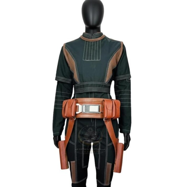 May include: A dark green long-sleeved jumpsuit featuring light brown paneling, vertical stitching, and a brown utility belt with two side pouches and a central metallic buckle. The outfit includes two long brown holsters hanging from the belt.