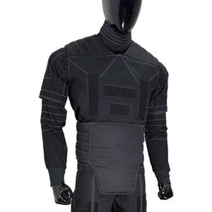 May include: Black tactical suit displayed on a mannequin. The suit features a high collar, long sleeves, and reinforced panels with white stitching. The design includes multiple Velcro patches and a padded lower torso section. The mannequin has a black, reflective head.