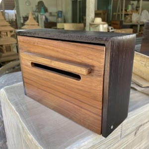 May include: A wooden mailbox, rectangular in shape, with a light brown front panel featuring a horizontal slot and a small handle. The top and sides are dark brown. Suitable for outdoor use.