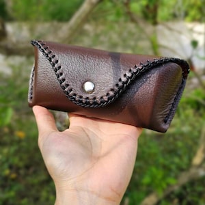 以下が含まれることがあります： ステッチされた黒い縁とシルバーのボタン留めが付いた、茶色の革製メガネケース。長方形のケースは手作りのようです。背景はぼやけており、屋外環境を示唆しています。