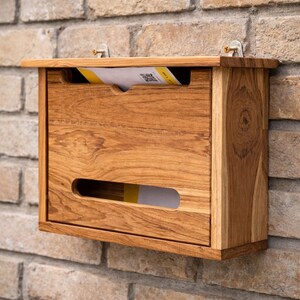 Könnte beinhalten: Ein Holzbriefkasten mit natürlicher Oberfläche ist an einer Ziegelmauer befestigt. Der Briefkasten hat eine rechteckige Form mit einem Schlitz für eingehende Post und einer unteren Öffnung zur Entnahme. Ein Brief ist im Inneren sichtbar.