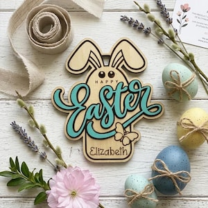 May include: A wooden Easter sign with a bunny silhouette and the words "Happy Easter" in teal script. The sign is personalized with the name "Elizabeth". The image also includes decorated Easter eggs, flowers, and ribbon.