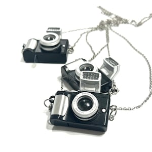 May include: Black camera pendant necklaces with silver-colored chains. The miniature cameras have silver accents and a lens that reads "ZHAOXIANGO OPTICALS 14-42mm". The necklaces are on a white background.
