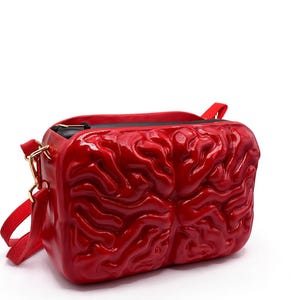 May include: A rectangular crossbody bag in a vibrant red colour featuring a three-dimensional, moulded brain-like texture on the front panel. The bag includes a black zippered top closure and a matching red adjustable shoulder strap with gold hardware.