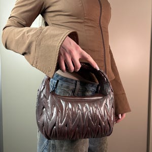 Puede incluir: Un bolso de mano en forma de media luna de color marrón oscuro, con una textura acolchada brillante y pliegues verticales fruncidos. El bolso cuenta con un asa corta integrada y una silueta redondeada, hecho de un material brillante.