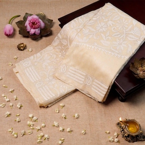 May include: Cream-coloured textile with intricate white floral embroidery. The fabric is folded and arranged on a brown surface, with small white flowers scattered around. Decorative elements include a pink flower, a brass dish, and a small oil lamp.