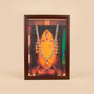 Puede incluir: Obra de arte enmarcada que presenta una deidad con rostro naranja, ojos grandes y un collar decorativo. La imagen está ambientada sobre un fondo oscuro con detalles verdes y marrones. El marco es de color marrón oscuro.
