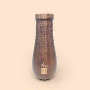 May include: A copper-toned water bottle with a hammered texture. The bottle has a rounded base and a tapered neck, with a matching lid. The words "isha LIFE" are printed in gold on the front of the bottle.