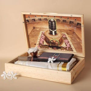 Op de afbeelding: Een houten kist met een scharnierend deksel, met religieuze items. Het deksel heeft een gedrukte afbeelding van een tempel met een centrale stenen structuur. Binnenin bevinden zich een kleine koperen vaas, een boek en een witte doek met gouden rand.