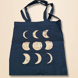 Puede incluir: Un bolso tote azul marino con un diseño de fases lunares doradas. El bolso tiene dos asas y está hecho de una tela resistente. Las fases lunares están dispuestas en un patrón circular, añadiendo un toque celestial al accesorio.